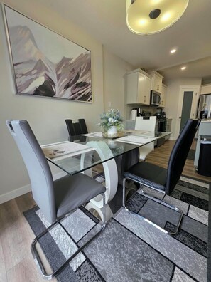 Un comedor moderno con una mesa de vidrio, sillas negras y blancas, un centro de mesa decorativo y una cocina al fondo.