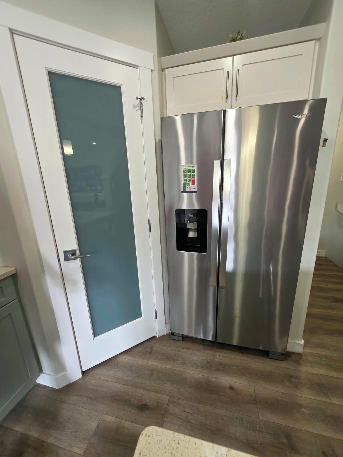 Un refrigerador de acero inoxidable con dispensador de agua, una puerta blanca con panel de vidrio esmerilado y gabinetes blancos.