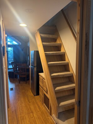 A staircase with carpeted steps leading to an upper floor, a wooden dining table with chairs, and a refrigerator in a kitchen area.