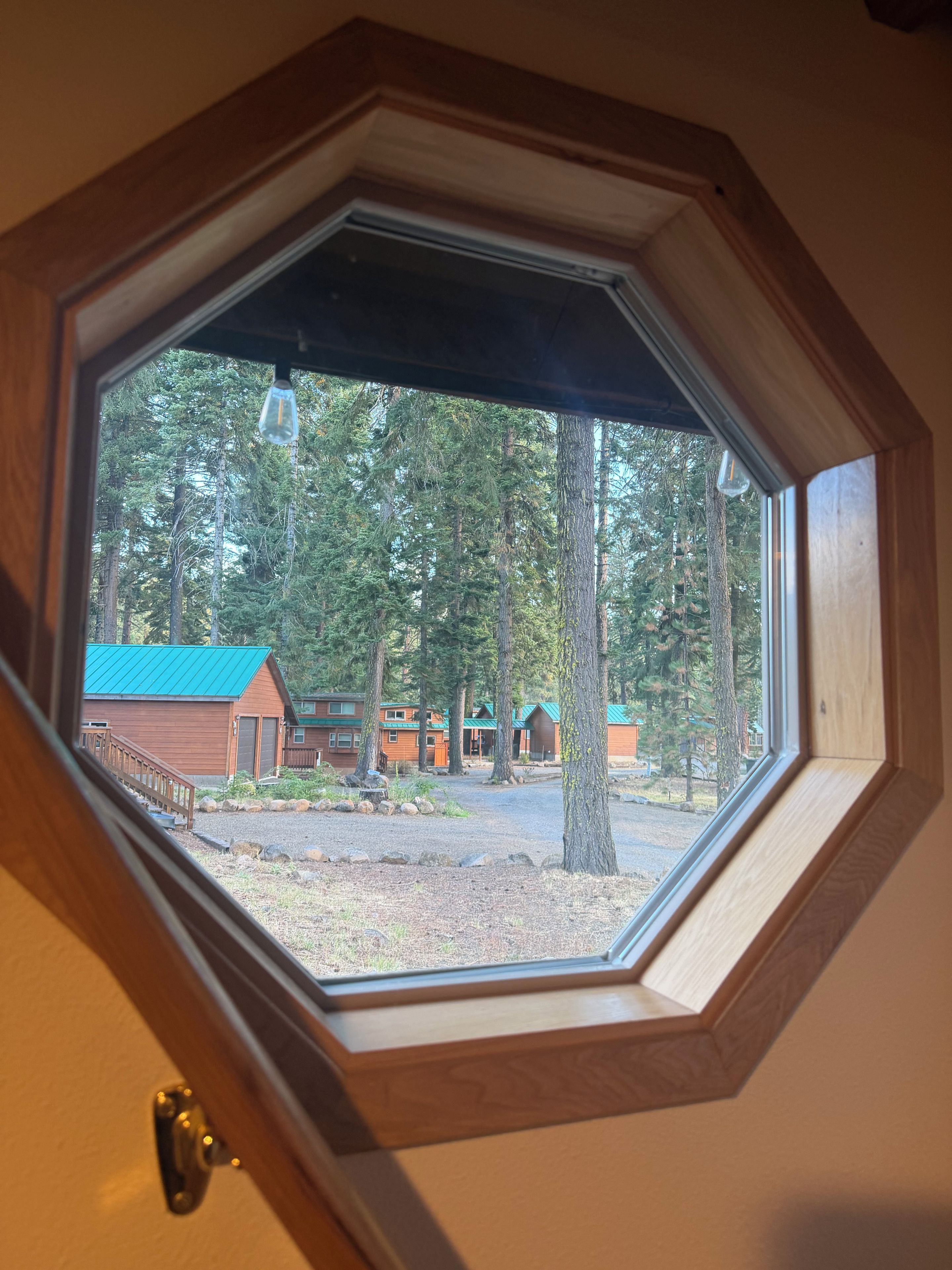Une vue à travers une fenêtre octogonale d’un chalet au toit vert, entouré d’arbres.