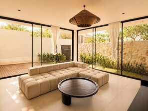 Un salon moderne avec un canapé d’angle, une table basse ronde et de grandes fenêtres donnant sur un jardin et un mur en pierre.