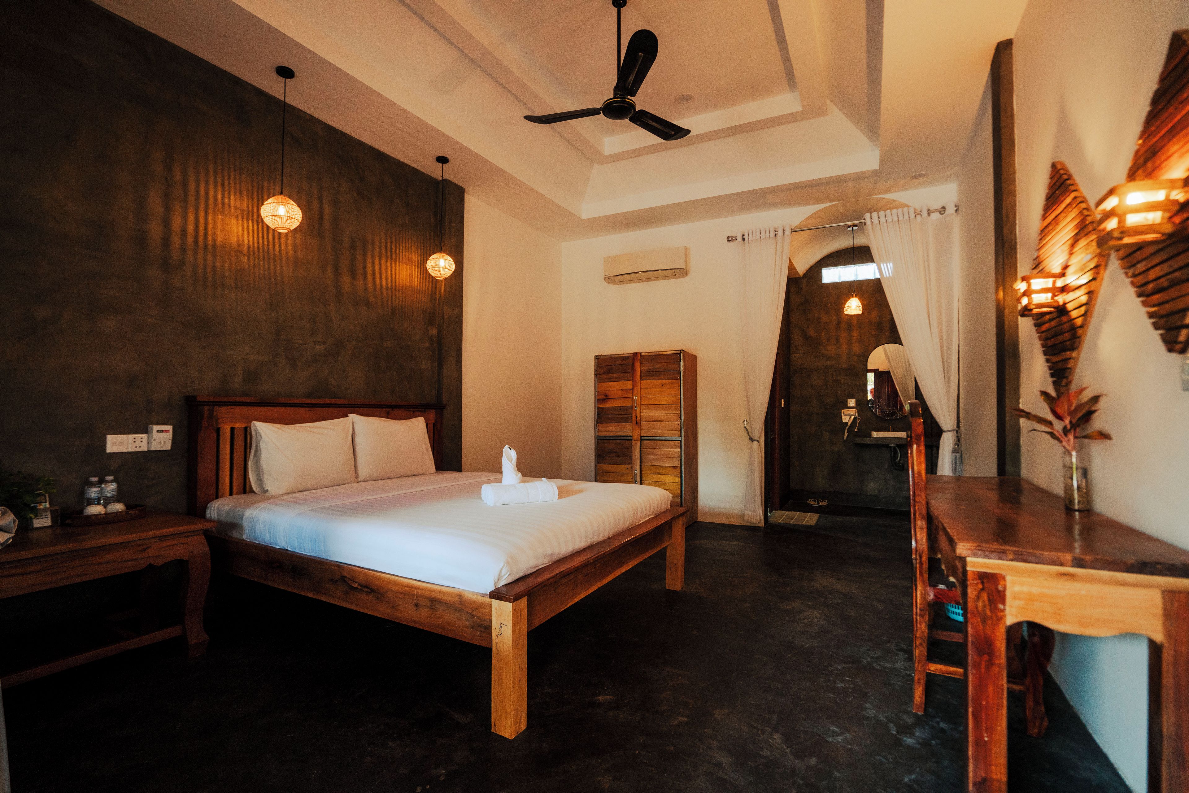 Quarto de hotel com cama de madeira, escrivaninha e ventoinha no teto.