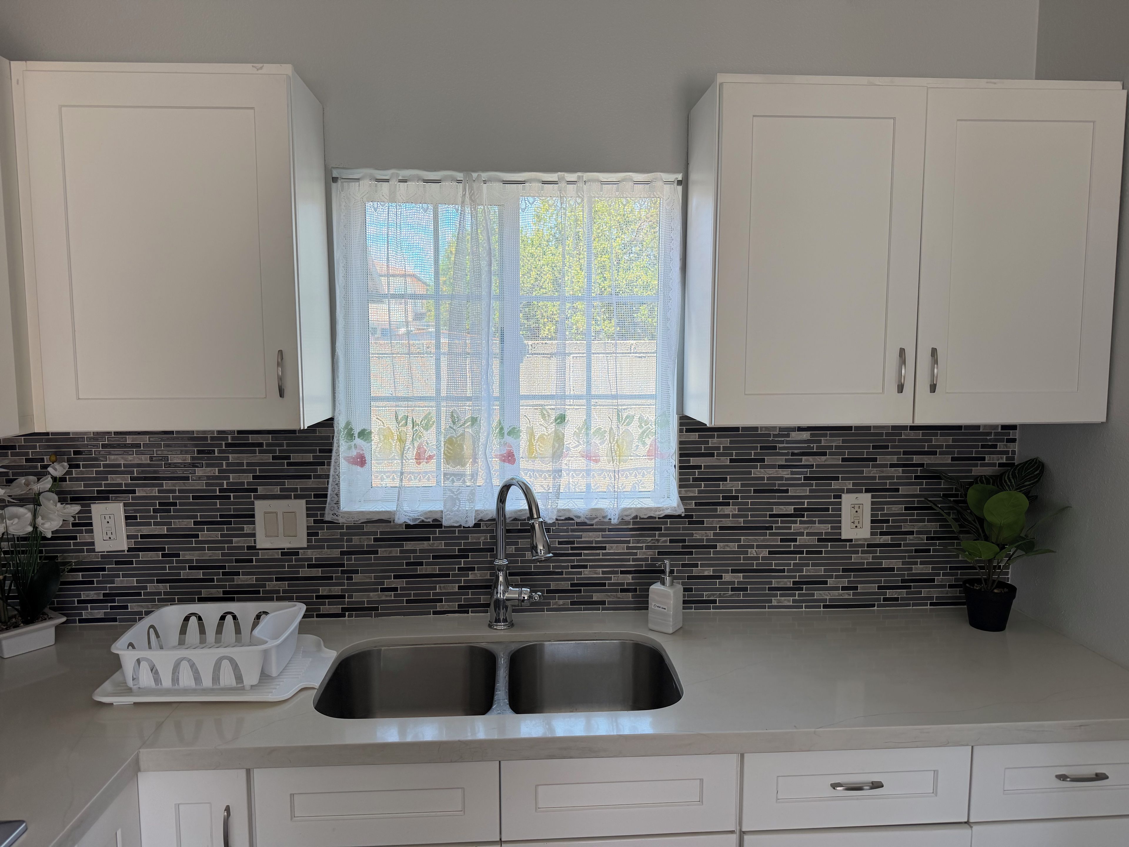Una cocina con dos lavamanos, armarios blancos, un ventanal con cortina de encaje y un salpicadero con azulejos estampados.