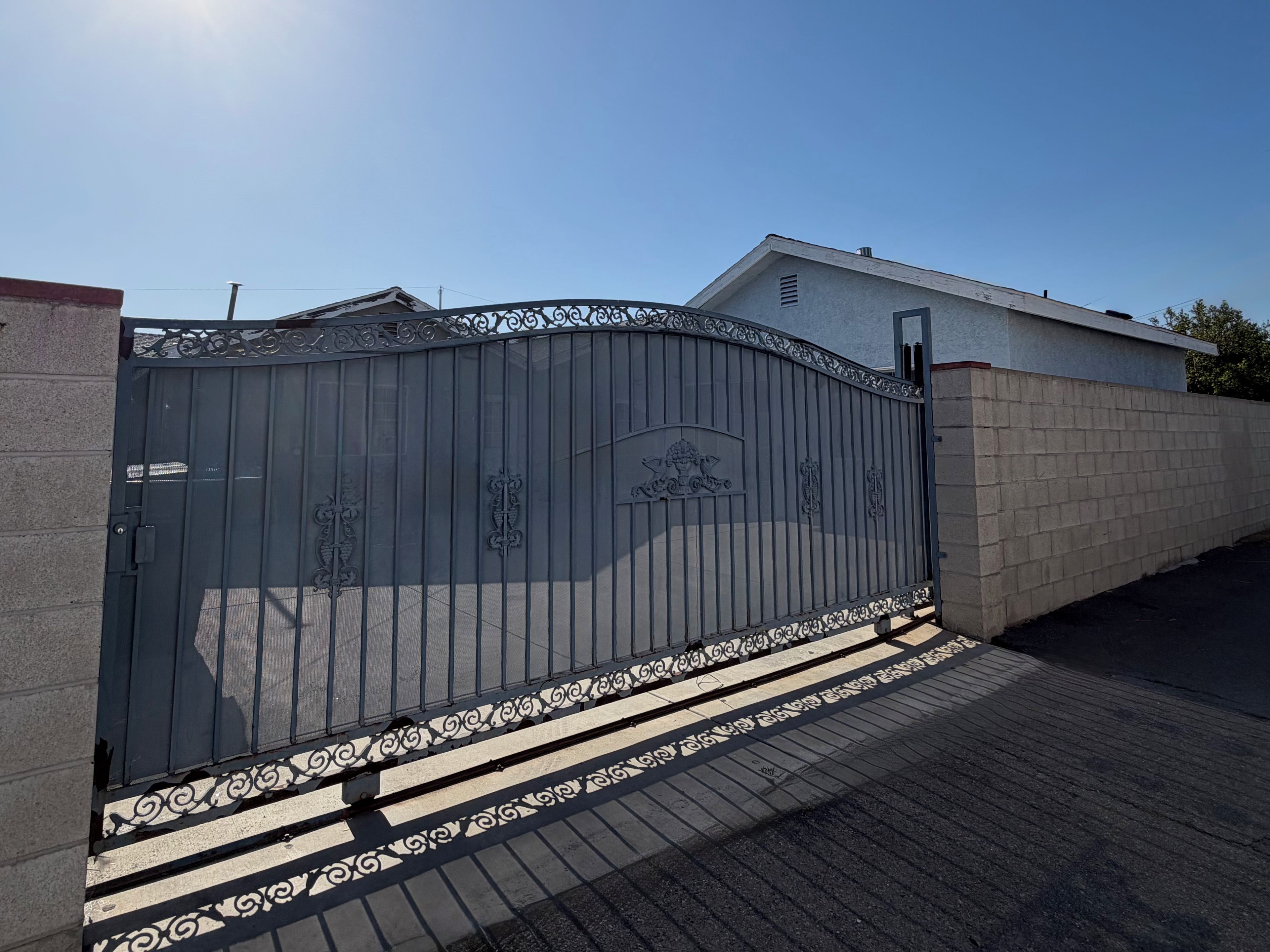 Una entrada protegida por un portón metálico decorativo, un muro de hormigón y un camino pavimentado que conduce a un edificio.