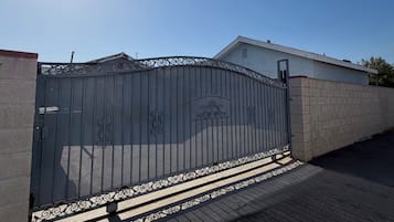Una entrada protegida por un portón metálico decorativo, un muro de hormigón y un camino pavimentado que conduce a un edificio.