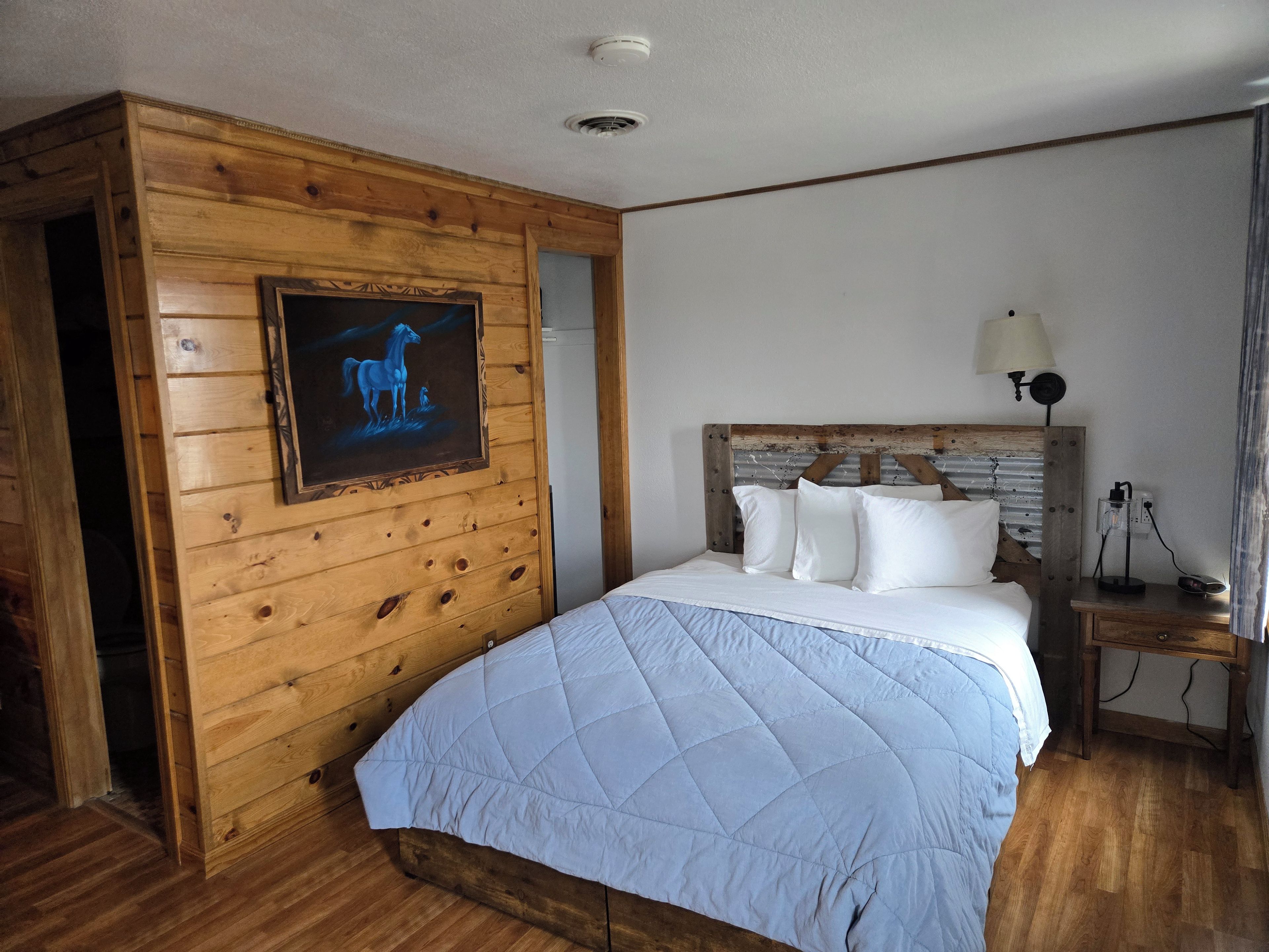 A room with a wooden headboard, a bed with white bedding, a nightstand with a lamp, and a wall-mounted picture of a horse.