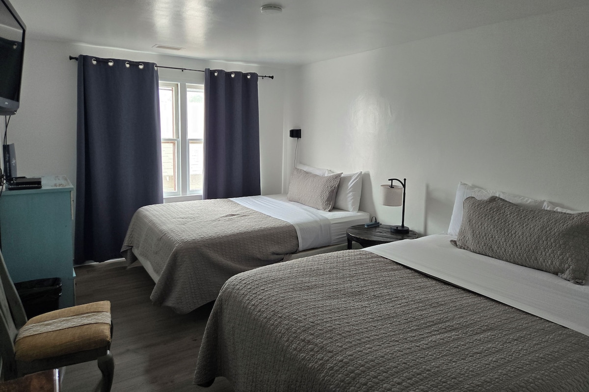 Habitació d'hotel amb dos llits, escriptori, cadira, làmpada i finestra amb cortines.