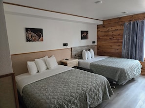 Quarto de hotel com duas camas, cabeceira de madeira, mesa de cabeceira e duas luminárias fixadas na parede.
