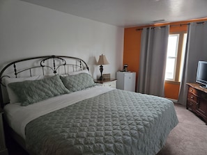 A bed with a quilted bedspread, a nightstand with a lamp, a small refrigerator, and a television on a wooden dresser.