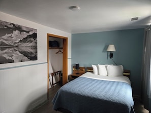 Quarto de hotel com cama, mesa de cabeceira, luminária, guarda-roupa e um quadro na parede.