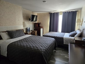 A hotel room with two beds, a flat-screen TV mounted on the wall, a desk with a lamp, and a window with curtains.
