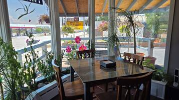 Un área de comedor con mesa y sillas de madera, ventanas amplias con vista a la calle y plantas en macetas.
