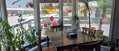 Un área de comedor con mesa y sillas de madera, ventanas amplias con vista a la calle y plantas en macetas.