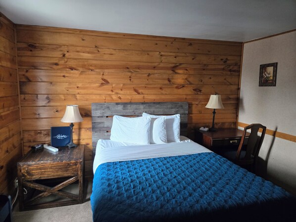 A hotel room with a wooden paneled wall, a bed with white linens, a blue quilted bedspread, two bedside lamps, and a small table with a lamp and a decorative box.