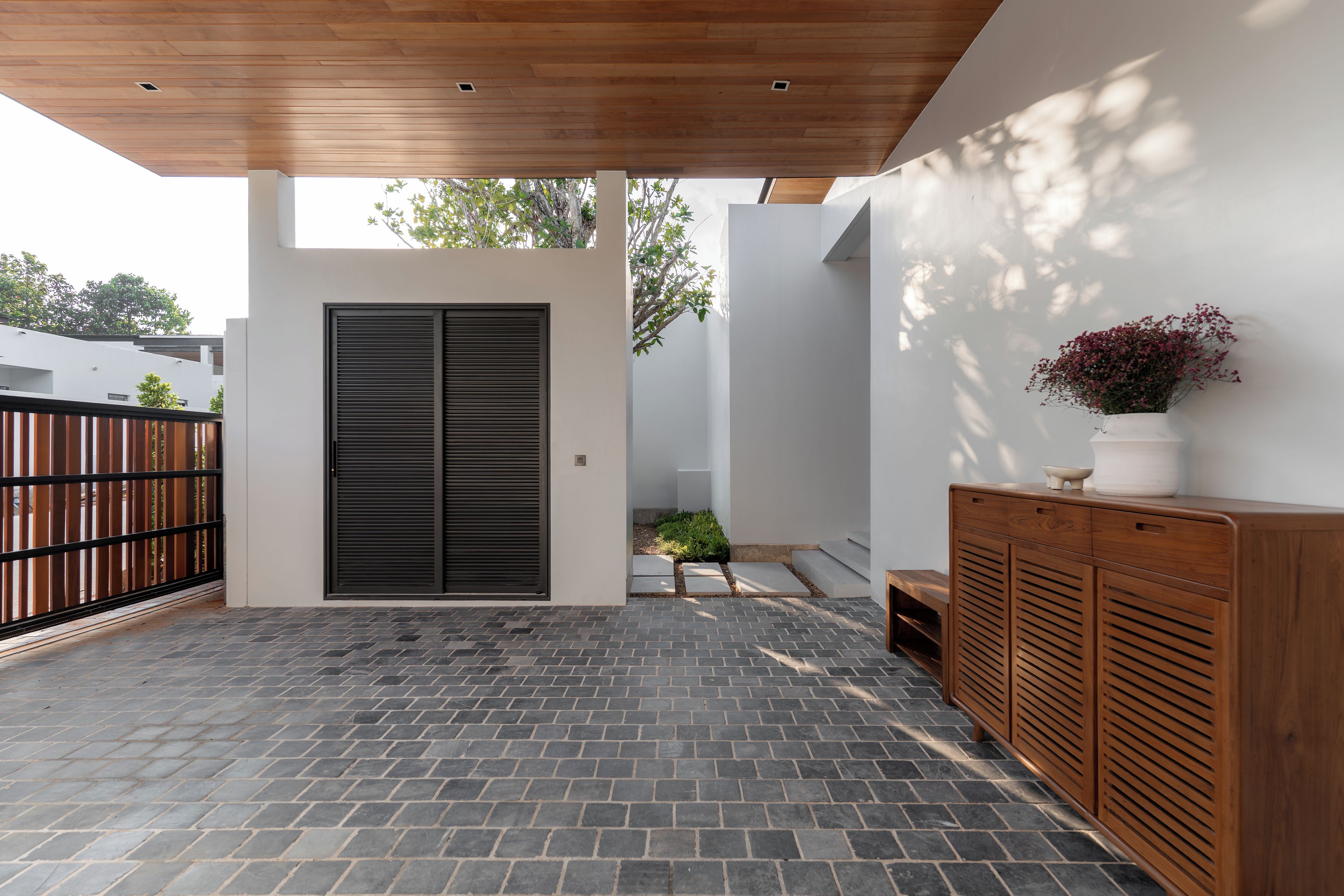 Une terrasse extérieure moderne avec un plafond en bois, une porte à volets fermés et un meuble en bois orné d’une plante en pot.