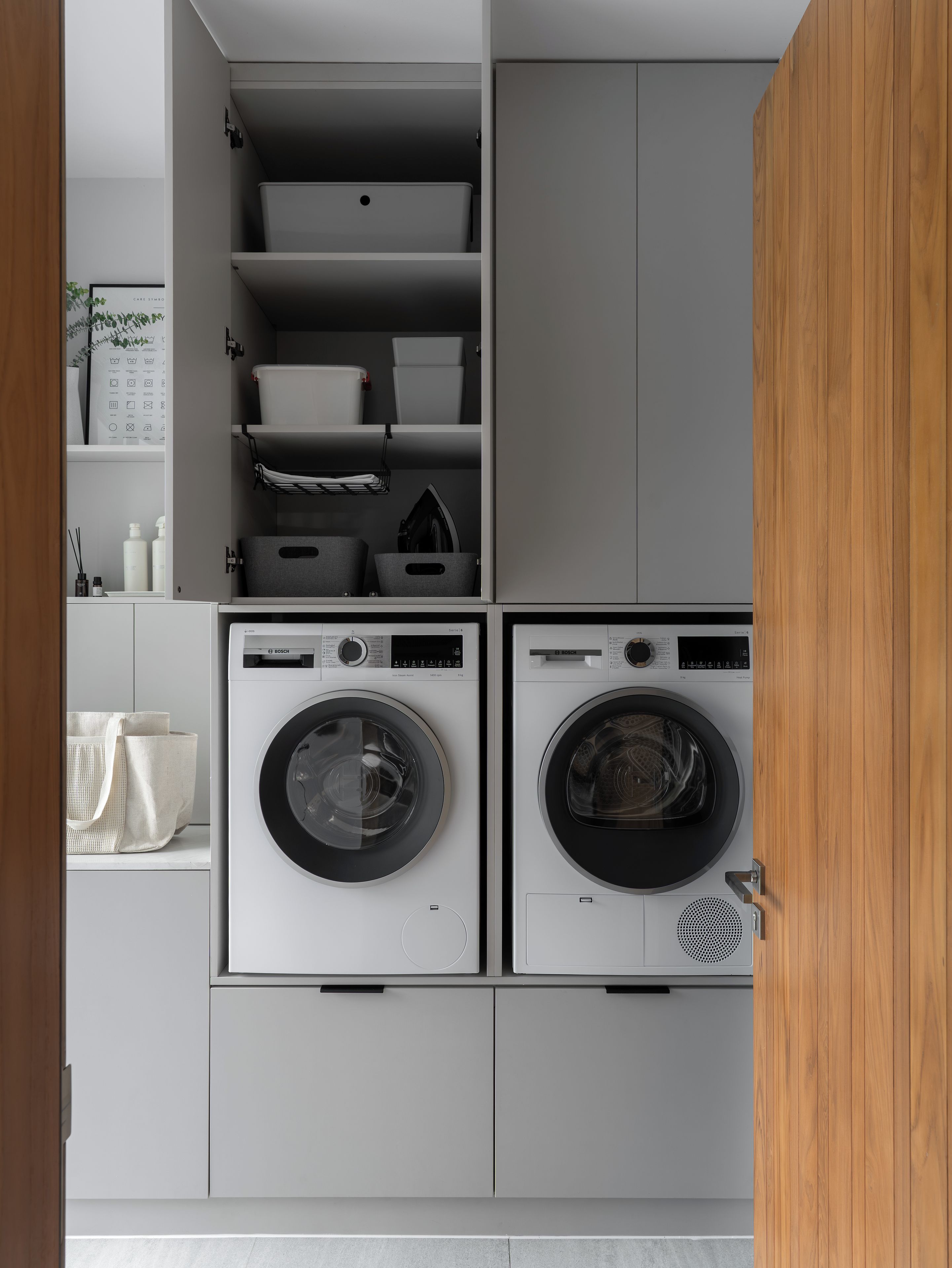 A laundry room with two front-loading washing machines, storage cabinets, and a shelf with various items.