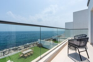En balkon med gelænder af glas, der kigger ud over havet, møbleret med stole og et bord.