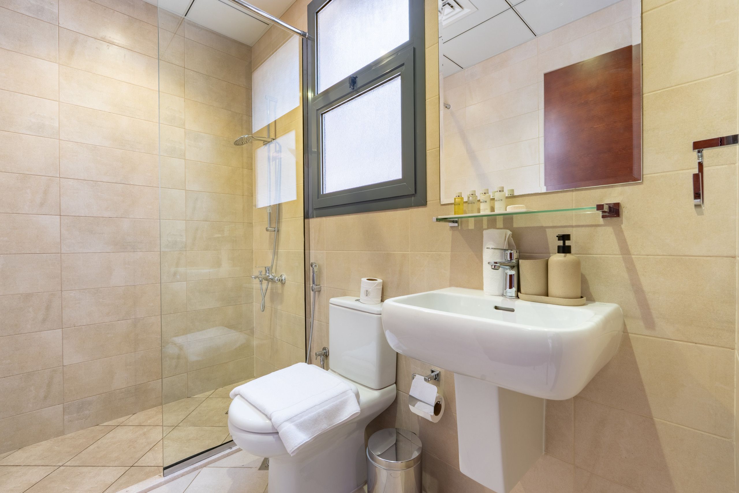 Casa de banho moderna com cabine de chuveiro em vidro, sanita branca, lavatório com espelho e artigos de higiene numa prateleira.