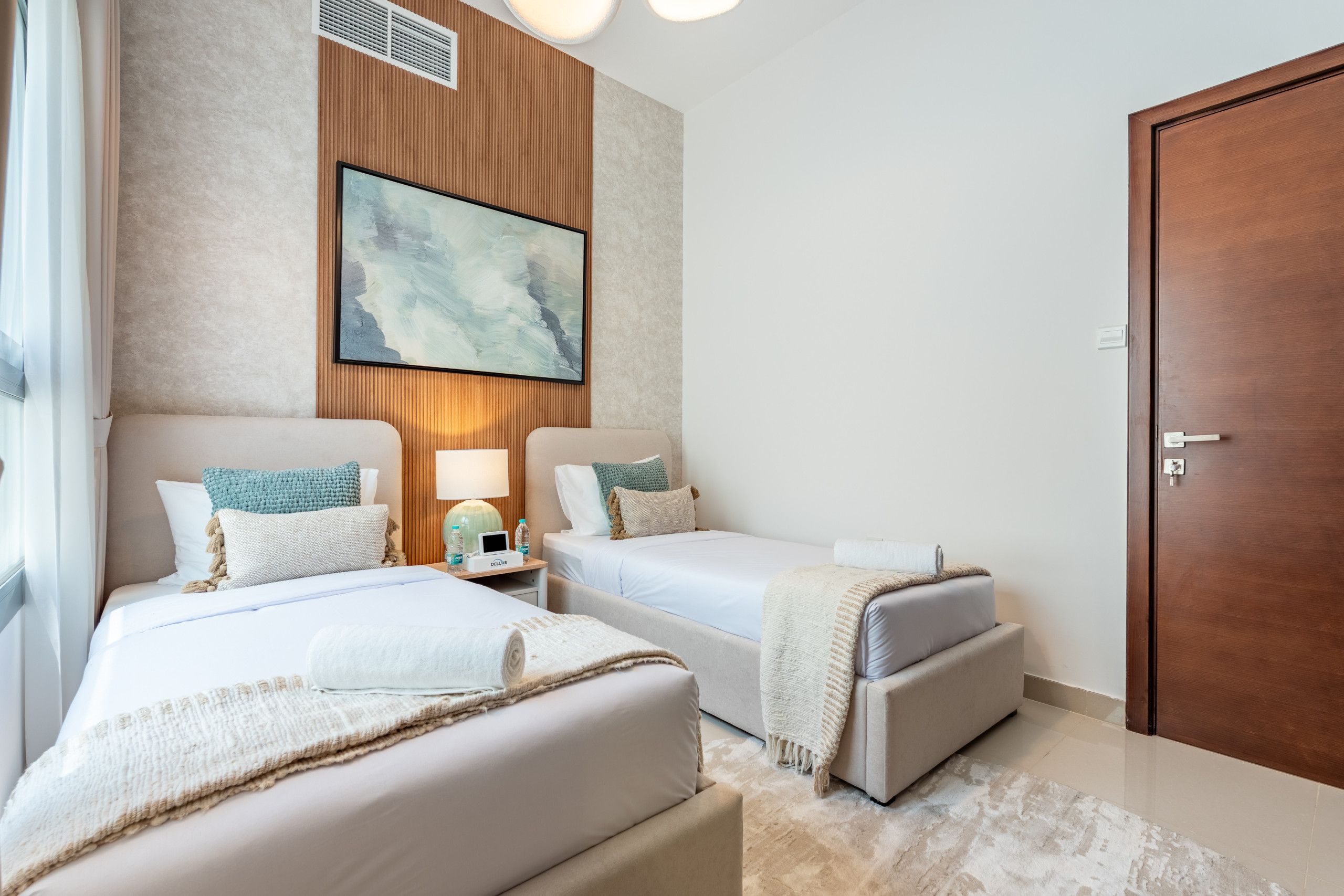 Habitación de hotel con dos camas, un cuadro abstracto grande, pared de paneles de madera, una puerta y una ventana con cortinas.