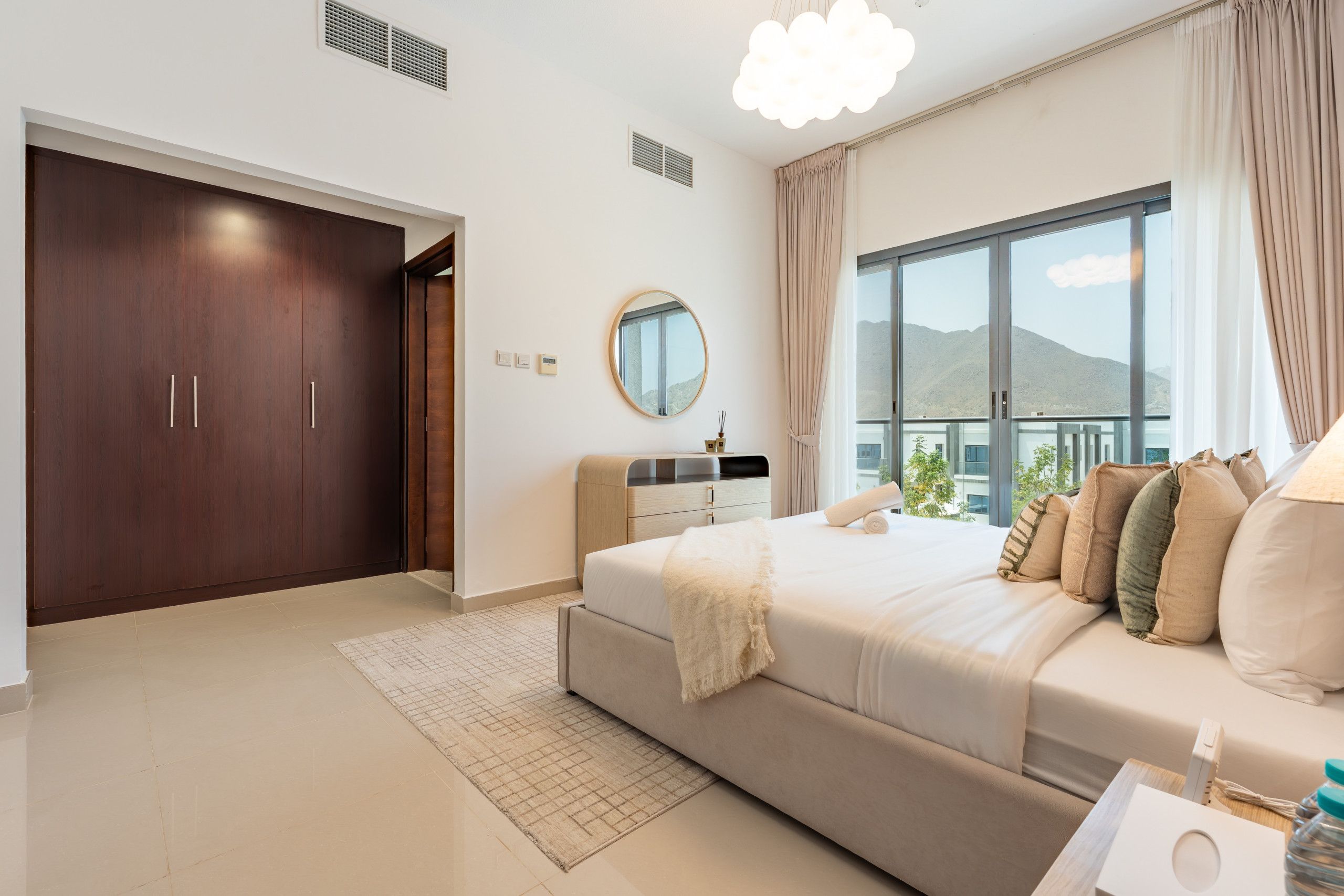 A modern bedroom with a large bed, a dresser, a mirror, and a window with a view of mountains.