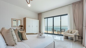 A modern bedroom with a large bed, a wardrobe, a seating area with a view of the sea, and a balcony. - Contemporary 4BR Villa with Assistant & Driver Rooms, Gulf Views, BBQ and Outdoor Lounge (Fujairah)
