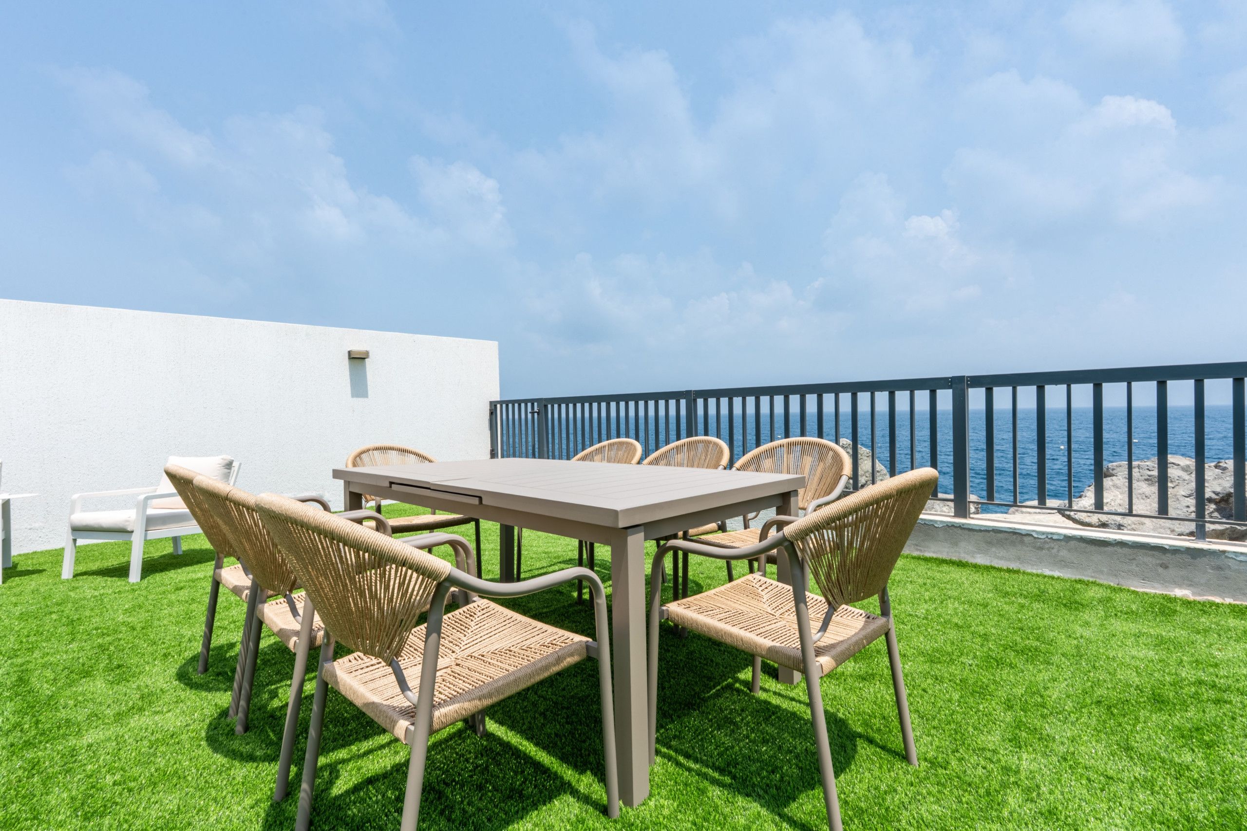 An outdoor patio with a table and chairs, a white wall, and a view of the sea.