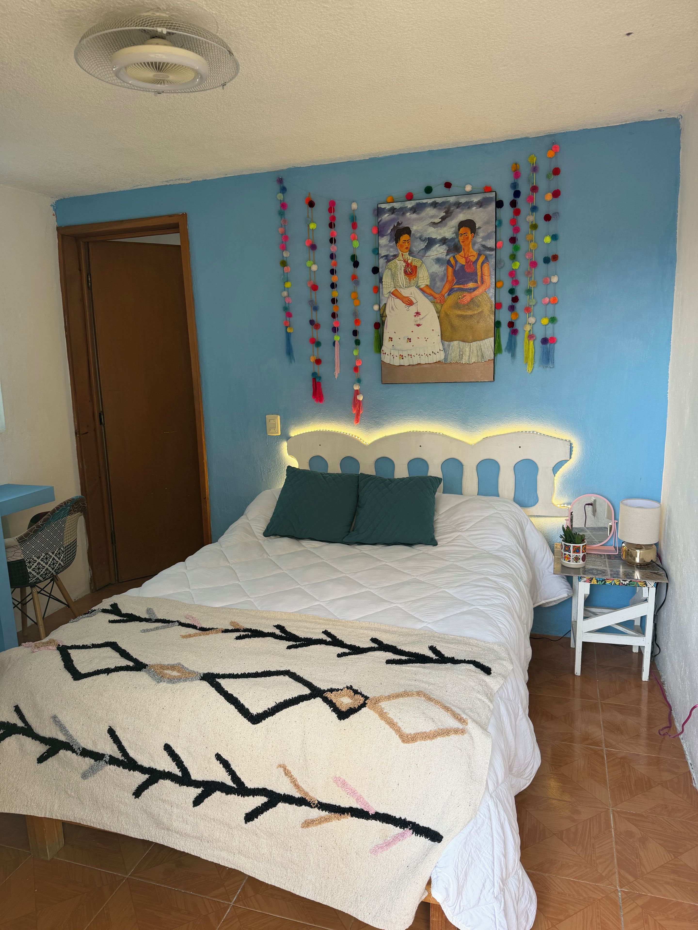 A bedroom with a bed, a bedside table, a chair, a wall painting, and a ceiling fan.
