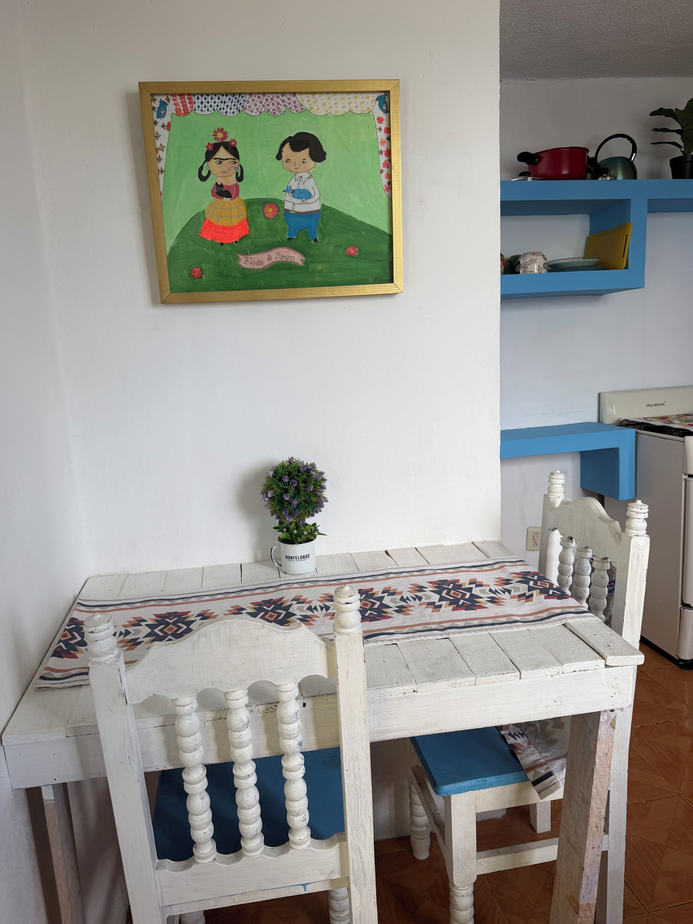 Un coin repas avec une table et des chaises, un tableau accroché au mur, une plante en pot et une étagère bleue contenant des ustensiles de cuisine.