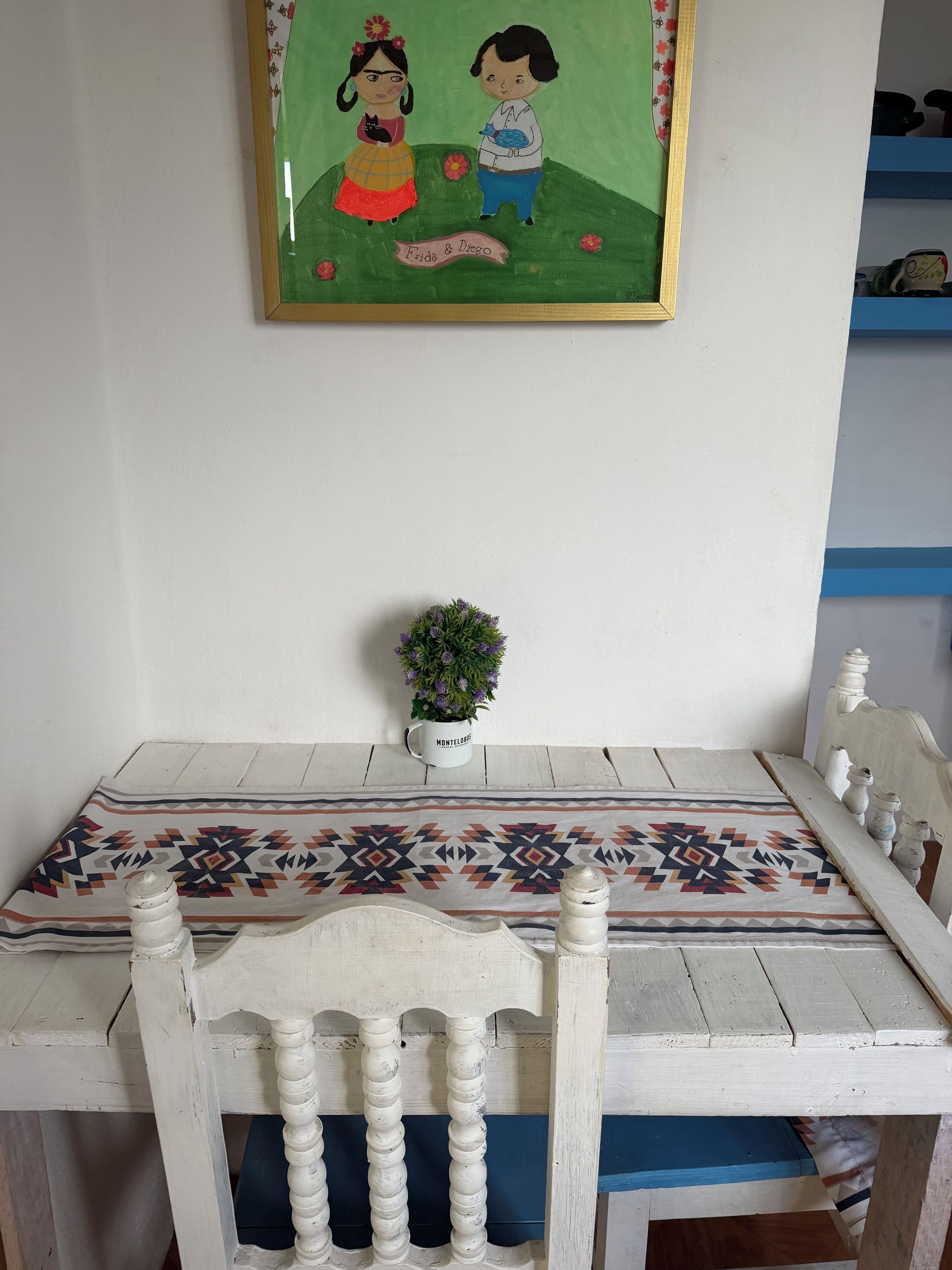 A dining table with a patterned tablecloth, white chairs, a small potted plant, and a framed artwork on the wall.