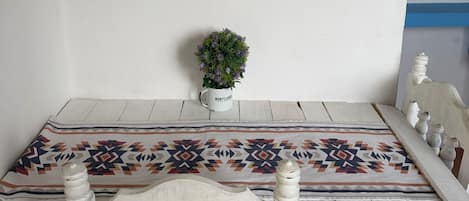 Uma mesa de jantar com toalha de mesa estampada, cadeiras brancas, um pequeno vaso com planta e uma obra de arte emoldurada na parede.