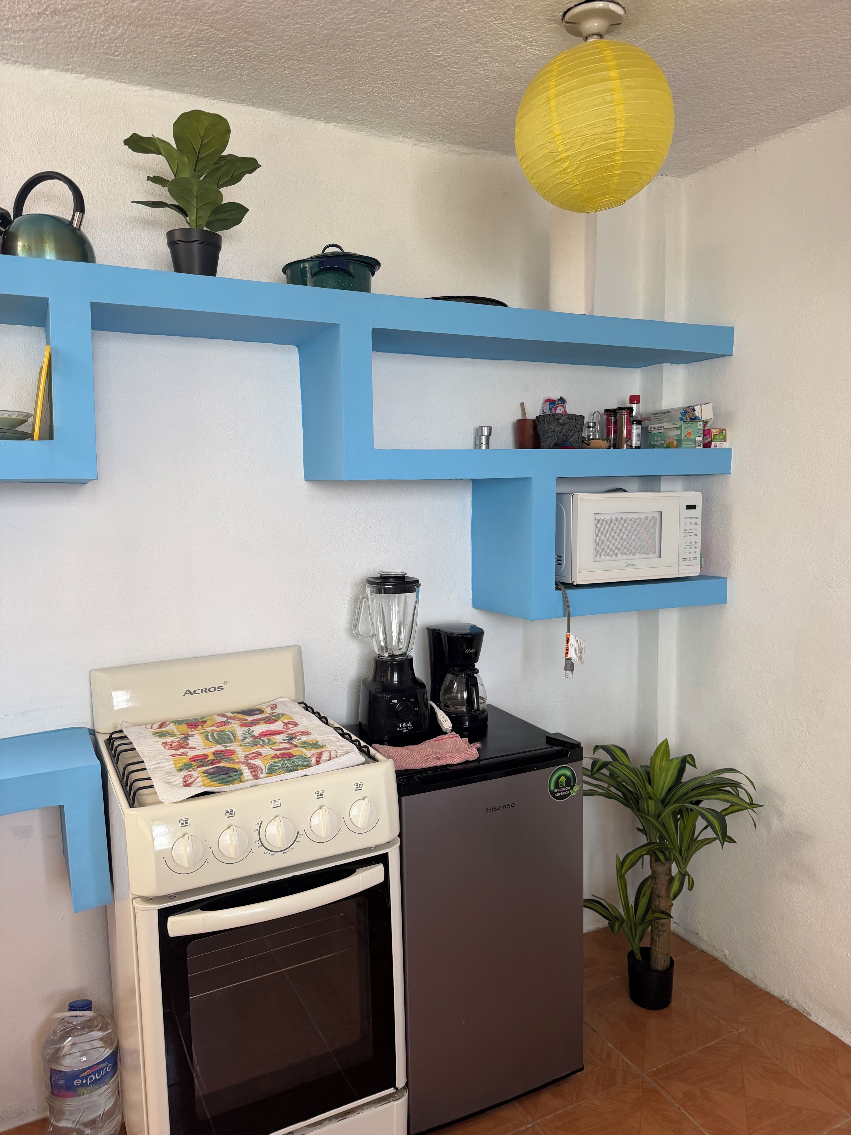 Una cocina compacta con estufa, microondas y licuadora sobre el mostrador. Hay repisas azules encima del mostrador con diversos objetos.