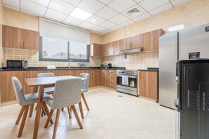 A modern kitchen with wooden cabinets, a dining table with chairs, a refrigerator, and a window with blinds.