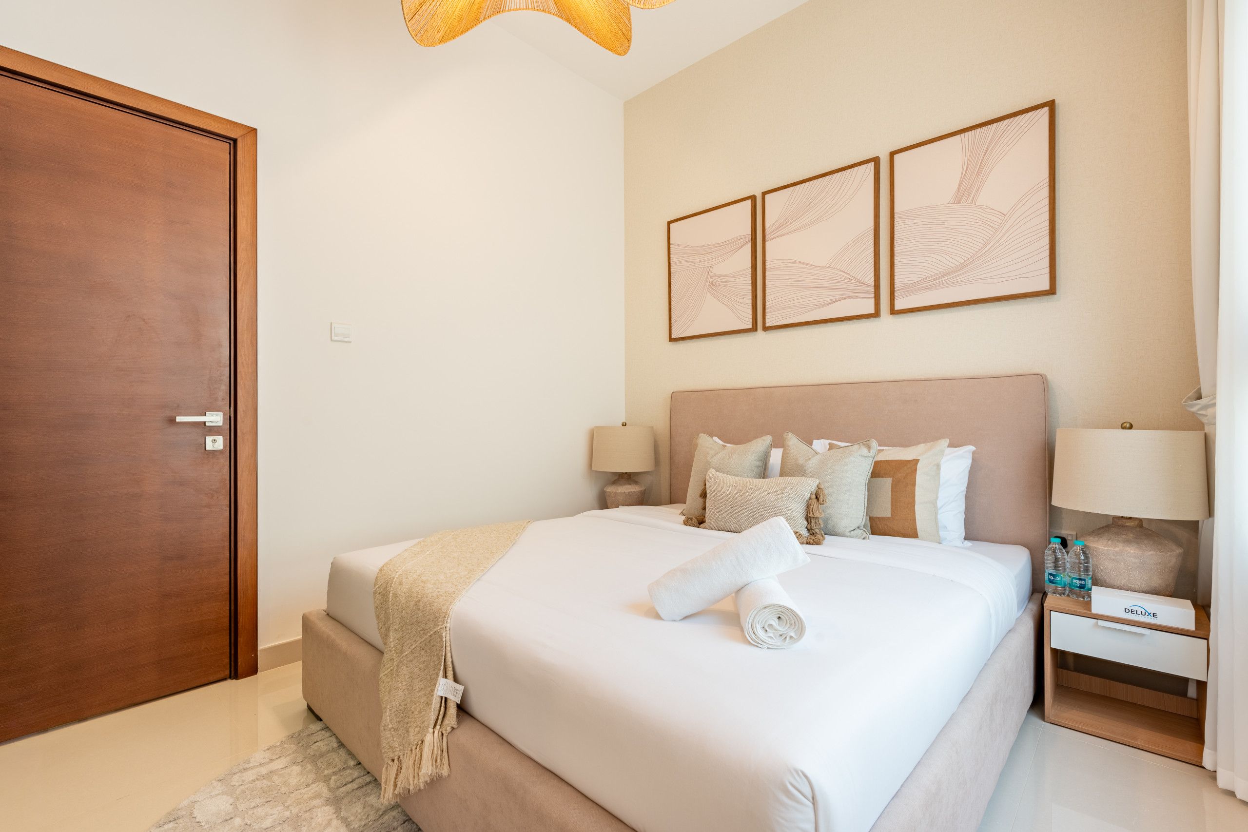 Habitación de hotel con una cama grande, lámparas de noche, una mesita de noche con una botella de agua y una puerta.