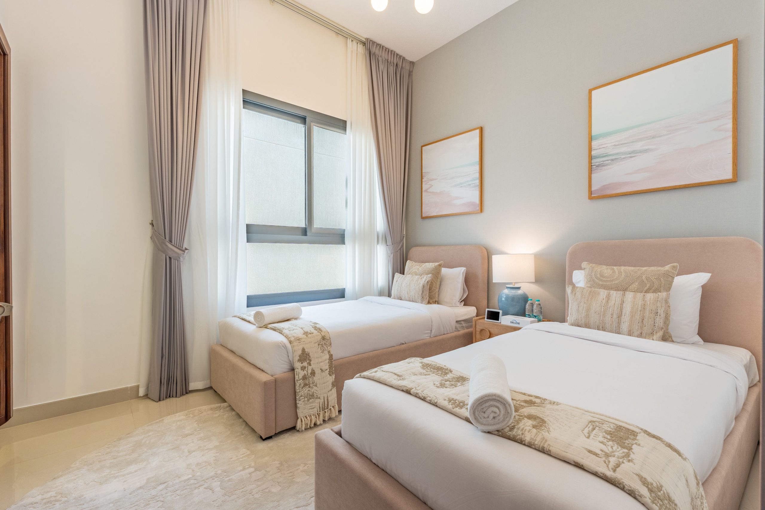 Quarto de hotel com duas camas, uma janela grande com cortinas, uma obra de arte pendurada na parede e uma mesa de cabeceira com abajur e garrafas de água.