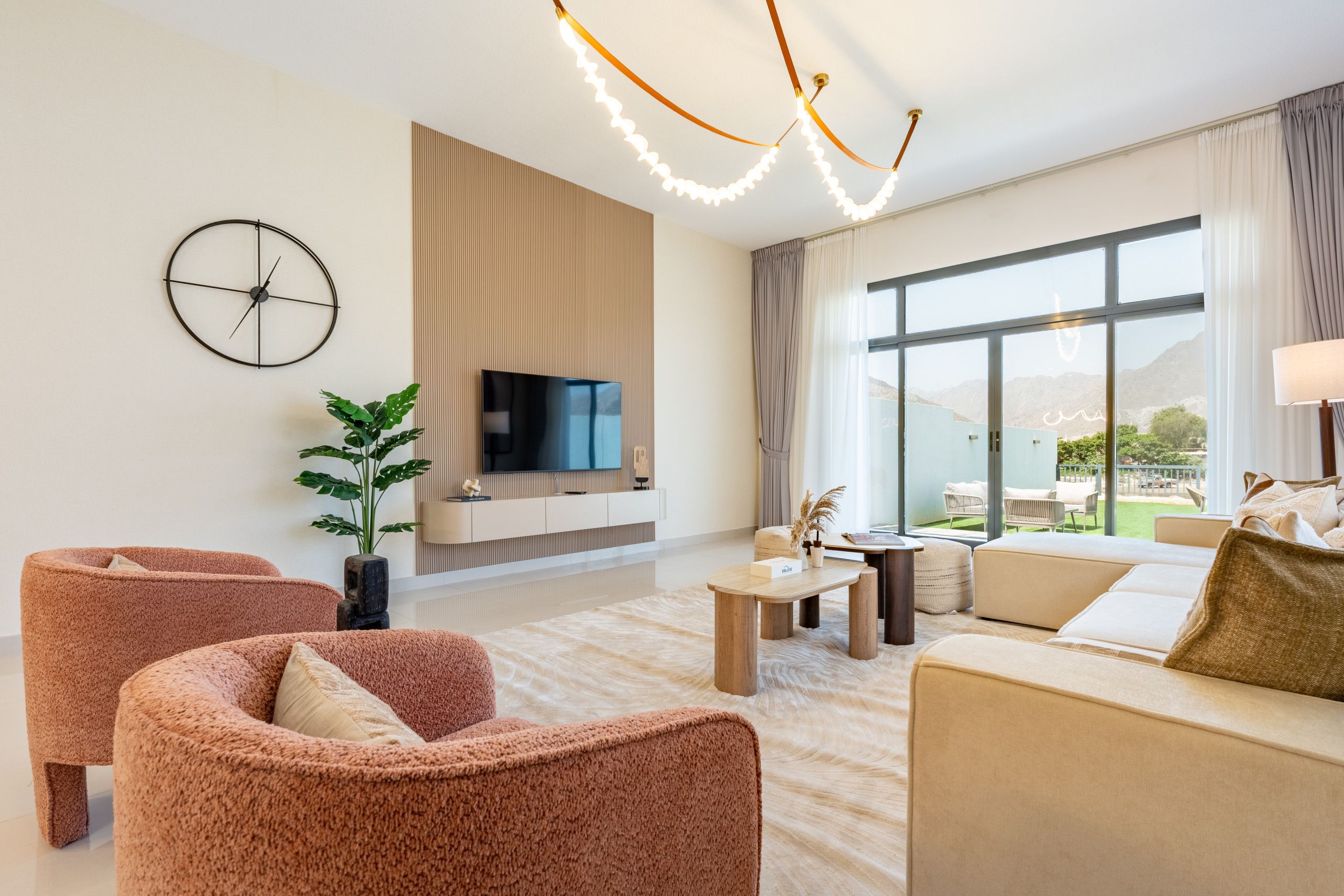 Una sala de estar moderna con una televisión de pantalla plana, dos sillones rosados, un sofá beige, una mesa de centro de madera y un ventanal con vistas al exterior.