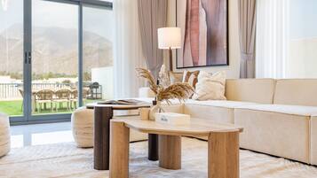 A modern living room with a sofa, a coffee table, and a lamp. There is a view of the outdoors through large windows.