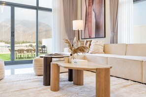 A modern living room with a sofa, a coffee table, and a lamp. There is a view of the outdoors through large windows.