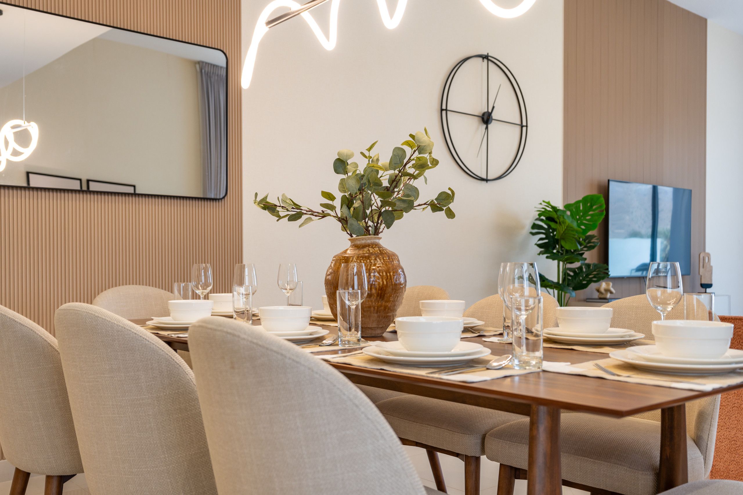 Un comedor con una mesa de madera dispuesta para una comida, rodeada de sillas, un jarrón grande con follaje y un reloj colgado en la pared.