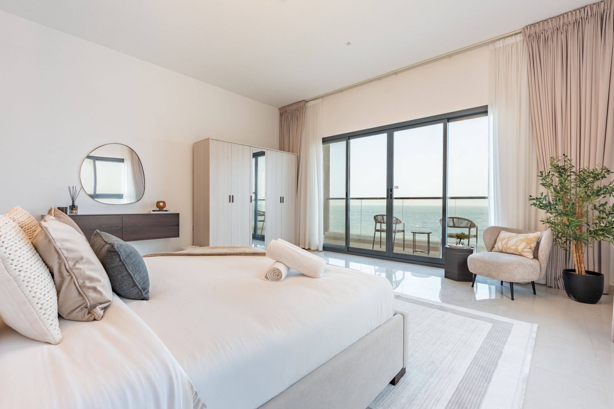 Un dormitorio moderno con una cama grande, un armario, un espejo y vistas al mar a través de ventanales de gran altura.