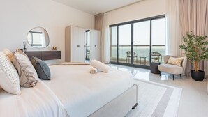Quarto moderno com uma cama grande, guarda-roupa, espelho e vista para o mar através de janelas do chão ao teto.