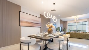 Uma área de jantar moderna com uma mesa comprida, cadeiras brancas e um vaso de flores secas.