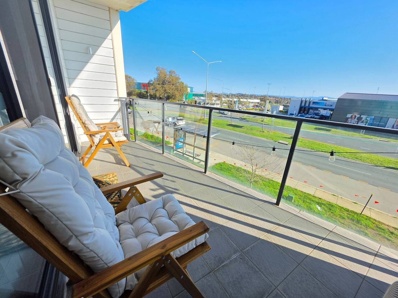 A balcony with a cushioned chair and a wooden chair, overlooking a road with vehicles and buildings in the distance.