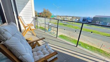 A balcony with a cushioned chair and a wooden chair, overlooking a road with vehicles and buildings in the distance.