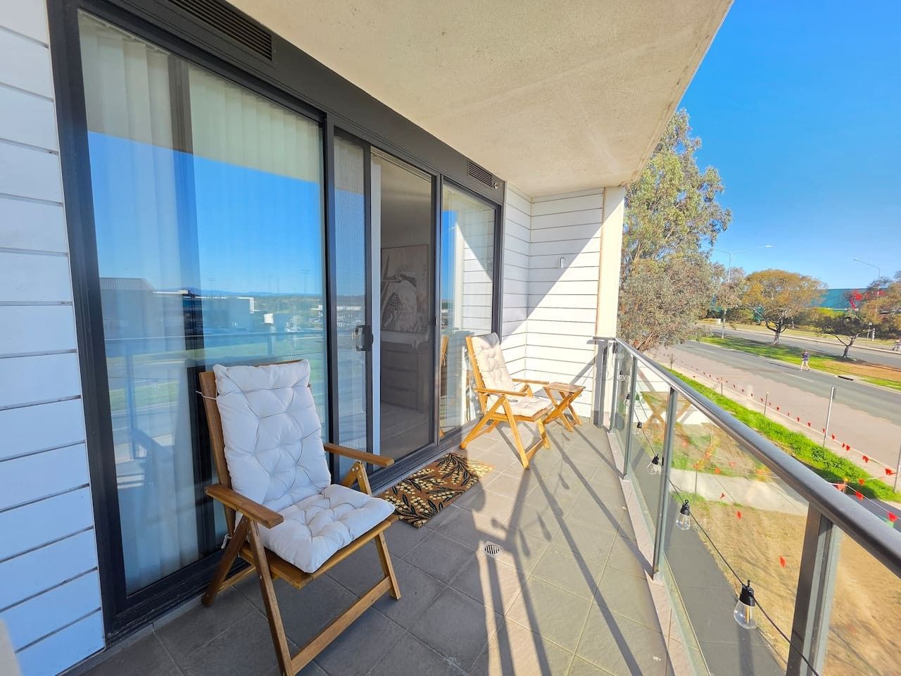 A balcony with a white cushioned chair and a wooden chair, a glass door, and a view of a road and trees.