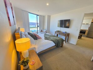 A hotel room with a bed, bedside table, lamp, TV, and a view of the city. - Gungahlin 1BR with Balcony, Pool, Gym and Spa (Gungahlin)