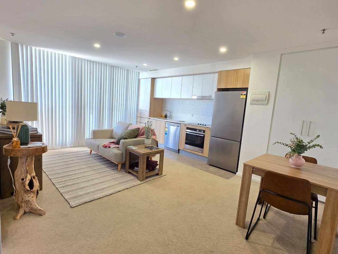 A modern living room with a sofa, coffee table, and a dining area with a table and chairs. A kitchen with a refrigerator and stove is visible in the background.