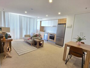 A modern living room with a sofa, coffee table, and a dining area with a table and chairs. A kitchen with a refrigerator and stove is visible in the background.