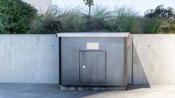 A concrete utility box with a door, located outdoors with a concrete wall and vegetation in the background.