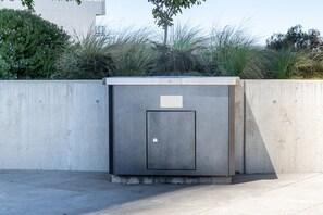 A concrete utility box with a door, located outdoors with a concrete wall and vegetation in the background.