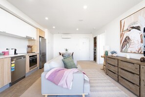 A modern kitchen with built-in appliances, a dining area with a table and chairs, and a living room with a sofa and a painting on the wall. - Gungahlin 1BR with Balcony, Pool, Gym and Spa (Gungahlin)